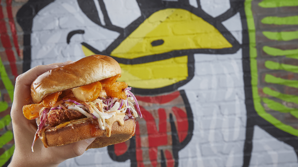 Handheld Bourbon Brown Sugar BBQ Sandwich with crispy chicken, tangy slaw, crunchy chips, and smoky BBQ sauce on a toasted bun.
